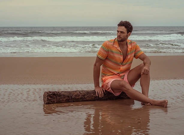 Homem vestindo uma camisa leve de linho na cor coral e shorts de praia, apreciando uma vista deslumbrante do litoral com falésias ao fundo e céu azul com nuvens esparsas. A combinação de roupa leve e elegante é ideal para o verão, trazendo conforto e estilo para momentos ao ar livre. Explore a coleção Código Zero, inspirada na natureza e no refúgio à beira-mar, com peças que unem sofisticação e funcionalidade. #camisalevedelinho #beachshortsdepraia #shortsmasculino #beachshorts #praia #litorabrasileiro #litoral #brasil #verao #codigozero