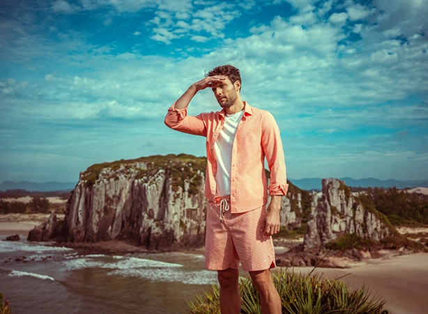Homem vestindo uma camisa leve de linho na cor coral e shorts de praia, apreciando uma vista deslumbrante do litoral com falésias ao fundo e céu azul com nuvens esparsas. A combinação de roupa leve e elegante é ideal para o verão, trazendo conforto e estilo para momentos ao ar livre. Explore a coleção Código Zero, inspirada na natureza e no refúgio à beira-mar, com peças que unem sofisticação e funcionalidade. #camisalevedelinho #beachshortsdepraia #shortsmasculino #beachshorts #praia #litorabrasileiro #litoral #brasil #verão #codigozero