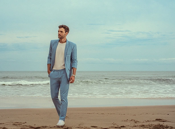Homem na praia, com mar ao fundo e céu, os dois em tons mais suaves e moderados. Ele veste uma camisa branca com tecido de alta qualidade, com blazer e calça em um azul claro e elegante. Na imagem conseguimos perceber que ele está caminhando com um semblante calma e relaxado. #alfaitaria #alfaitariamasculina #blazer #modapraia #praia #alfaitarialeve #leveza #alfaitariaveranil