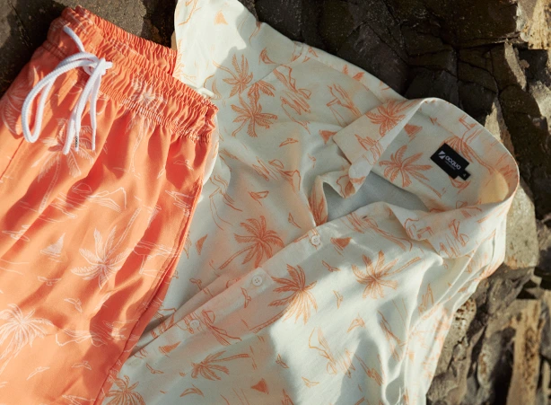 Bermuda beach shorts e camisa com estampas de coqueiros. Estão sobre uma pedra no qual é percebido que o sol está batendo trazendo uma sensação de tropicalidade a foto. #codigozero #beachshorts #camisaestampada #verao