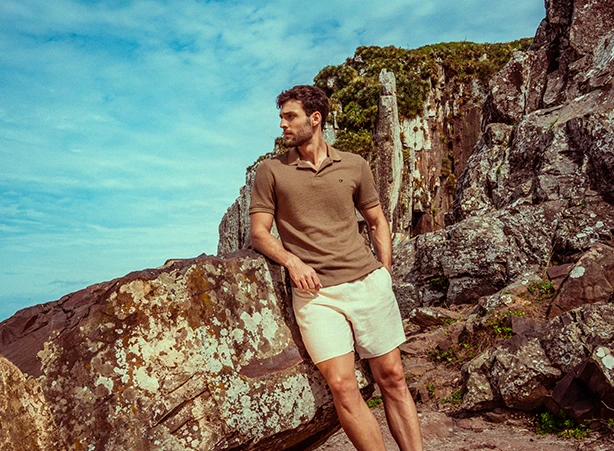 Homem de pé ao lado de rochas, em um cenário natural. Ele veste uma camisa polo de cor marrom-terra e bermuda clara de linho, criando um visual casual e sofisticado, ideal para ocasiões descontraídas ao ar livre. O homem usa óculos de sol com armação verde, que complementam o estilo leve e natural do look. A paleta de cores da roupa harmoniza com o ambiente rochoso, reforçando uma conexão com elementos da natureza e tons neutros. #lookveraomasculino #modapraiamasculino #texturasnaturais #estampas #conjuntomasculinoparapraia #estiloleve #polotexturizada