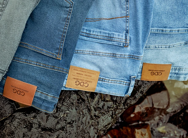Três calças jeans penduradas sobre algum elemento que parece ser uma rocha. As três calças são na cor azul denim mas em tonalidades diferentes, e apresentam uma etiqueta na cor caramelo identificando a marca da peça CDG da Código Zero. #jeans #jeansmasculino #lavanderiajeans #jeansparaoverao #denimmasculino #reinventandoojeans
