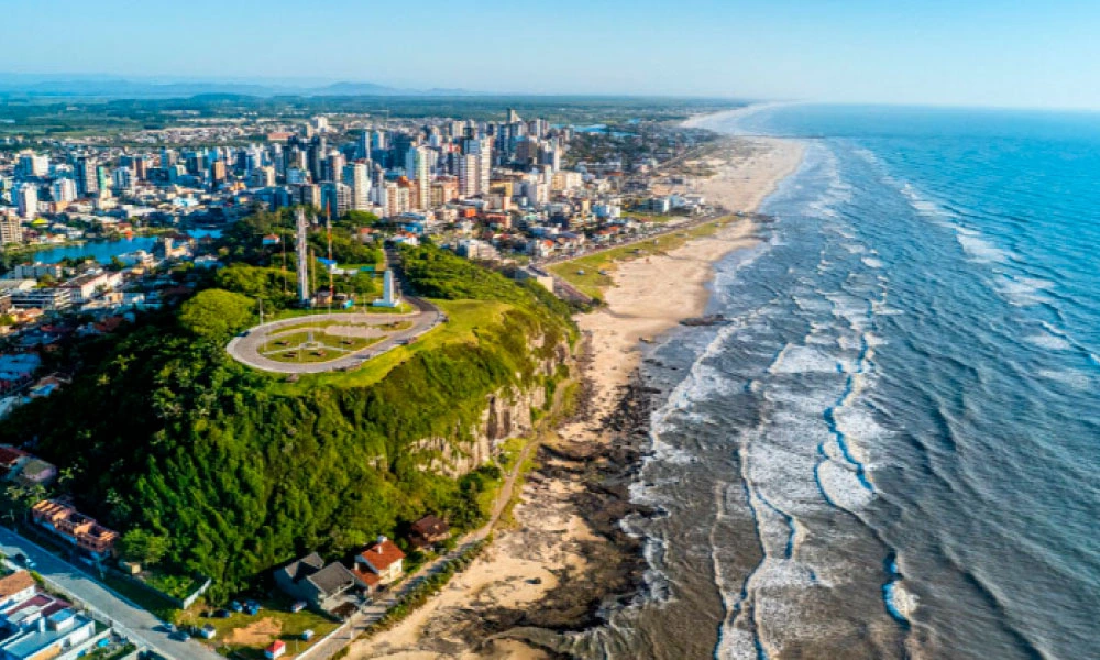 A imagem mostra a faixa litorânea de Torres-RS, com um contraste bonito entre os tons de verde do morro, o azul do mar e o desenvolvimento urbano com prédios e casas próximas à orla. Essa composição combina natureza e urbanismo, emoldurada pela costa marcante da região. #viagem #verao25 #codigozero #guiademala #viagemfimdeano #lookfinaldeano #praia #torres 
