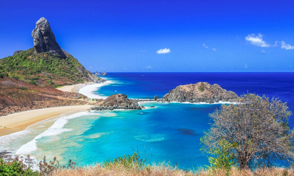 Na imagem observamos um dos mais famosos destinos litorâneos do Brasil. As praias de Fernando de Noronha são famosas por suas formações rochosas, que próximo do mar com azul cristalino forma uma paisagem única e deslumbrante. O céu azul e a vegetação em volta completam a imagem e a paisagem #viagem #verao25 #codigozero #guiademala #viagemfimdeano #lookfinaldeano #praia #fernandodenoronha 