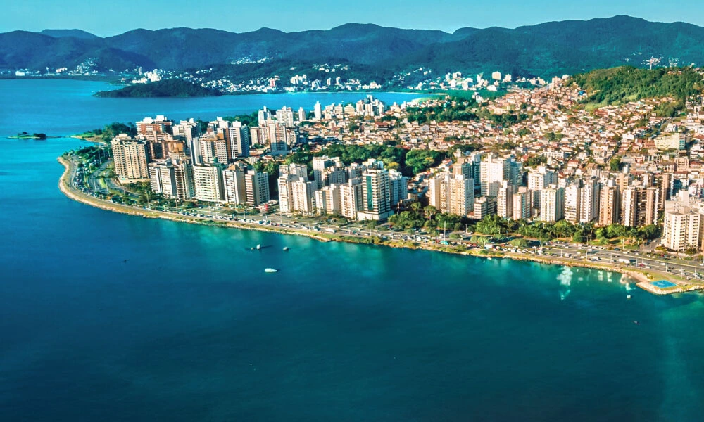 : A imagem mostra cidade de Florianópolis em Santa Catarina que realça de forma bem direta a parte litorânea Beira-mar Norte da cidade. É possível ver uma grande quantidade de prédios e ao fundo montanhas, assim como o mar azul contornando a cidade. Tags: #viagem #verao25 #codigozero #guiademala #viagemfimdeano #lookfinaldeano #praia #floripa 