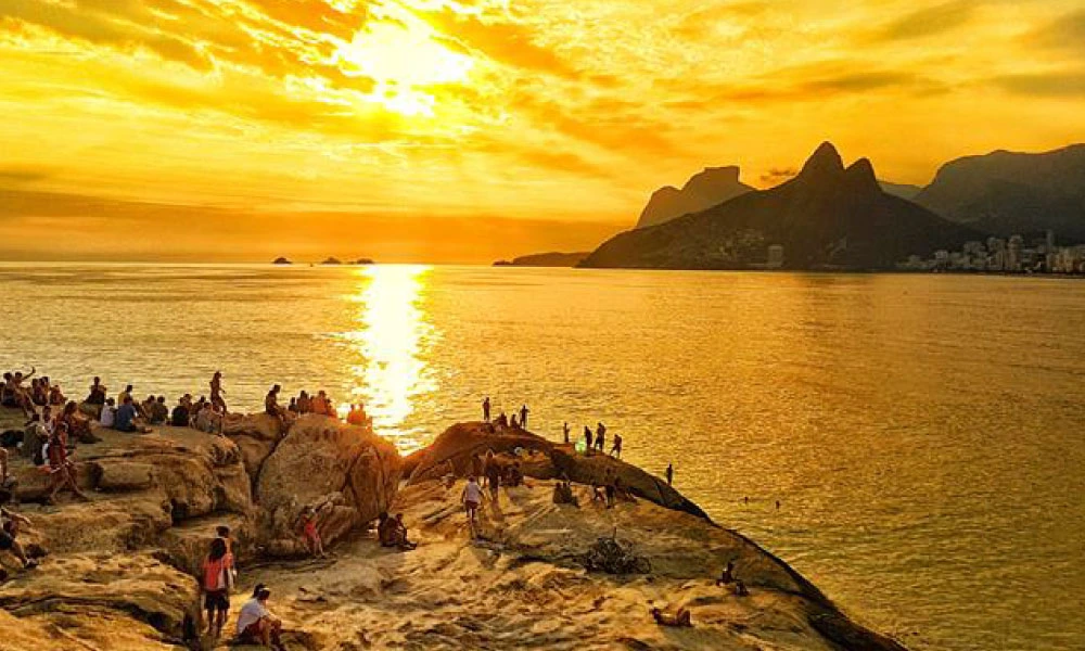 A imagem retrata um dos cenários mais emblemáticos do Rio de Janeiro, o pôr do sol visto da Pedra do Arpoador. O céu está tingido de tons dourados e alaranjados, refletindo no mar calmo, criando um espetáculo natural de luz. No horizonte, é possível identificar o contorno das montanhas, incluindo o Morro Dois Irmãos, que complementam a beleza da paisagem. Várias pessoas estão reunidas sobre as pedras, algumas sentadas e outras de pé, aproveitando o momento para contemplar a vista, socializar ou tirar fotos. A cena transmite um clima descontraído e típico de fim de tarde carioca, destacando a conexão das pessoas com a natureza e a cultura local. #viagem #verao25 #codigozero #guiademala #viagemfimdeano #lookfinaldeano #praia #riodejaneiro #arpoador 